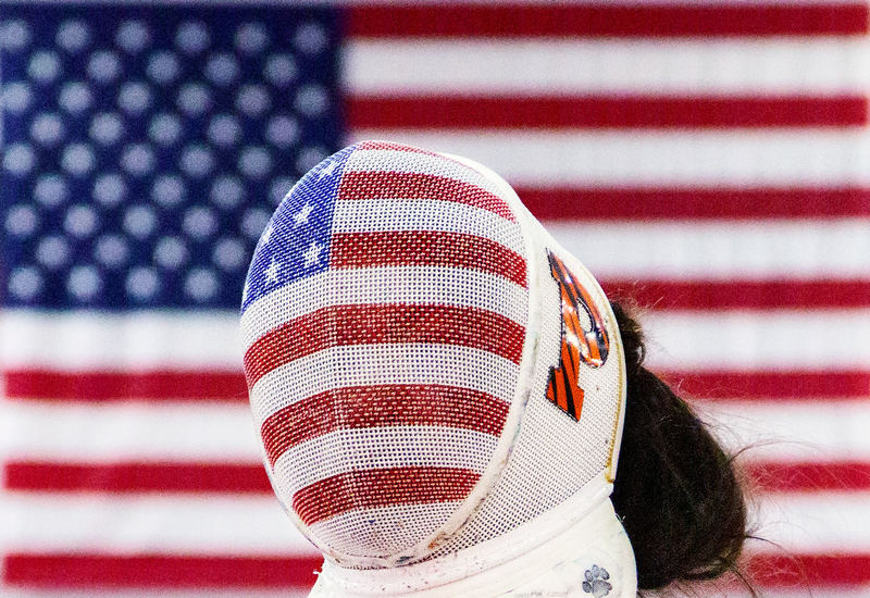 A fencer in a mask painted like the American flag, standing in front of a hanging flag