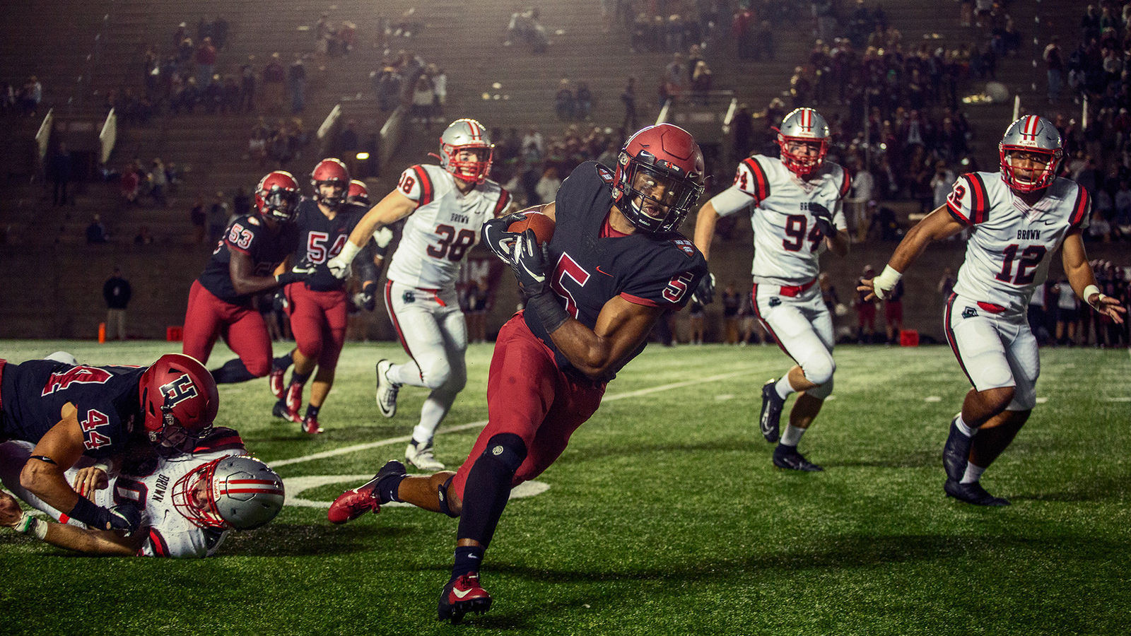 A running back in a dark number 5 jersey breaks a tackle under the lights